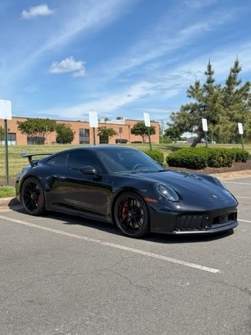 Black Porsche 911 sports car parked in a lot with brick buildings and landscaping in background