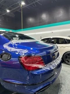 Blue sedan with protective plastic wrap on rear bumper at indoor car show or dealership with other vehicles visible