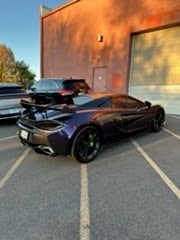 Black sports car parked in front of a large rust-colored metal structure in a parking lot